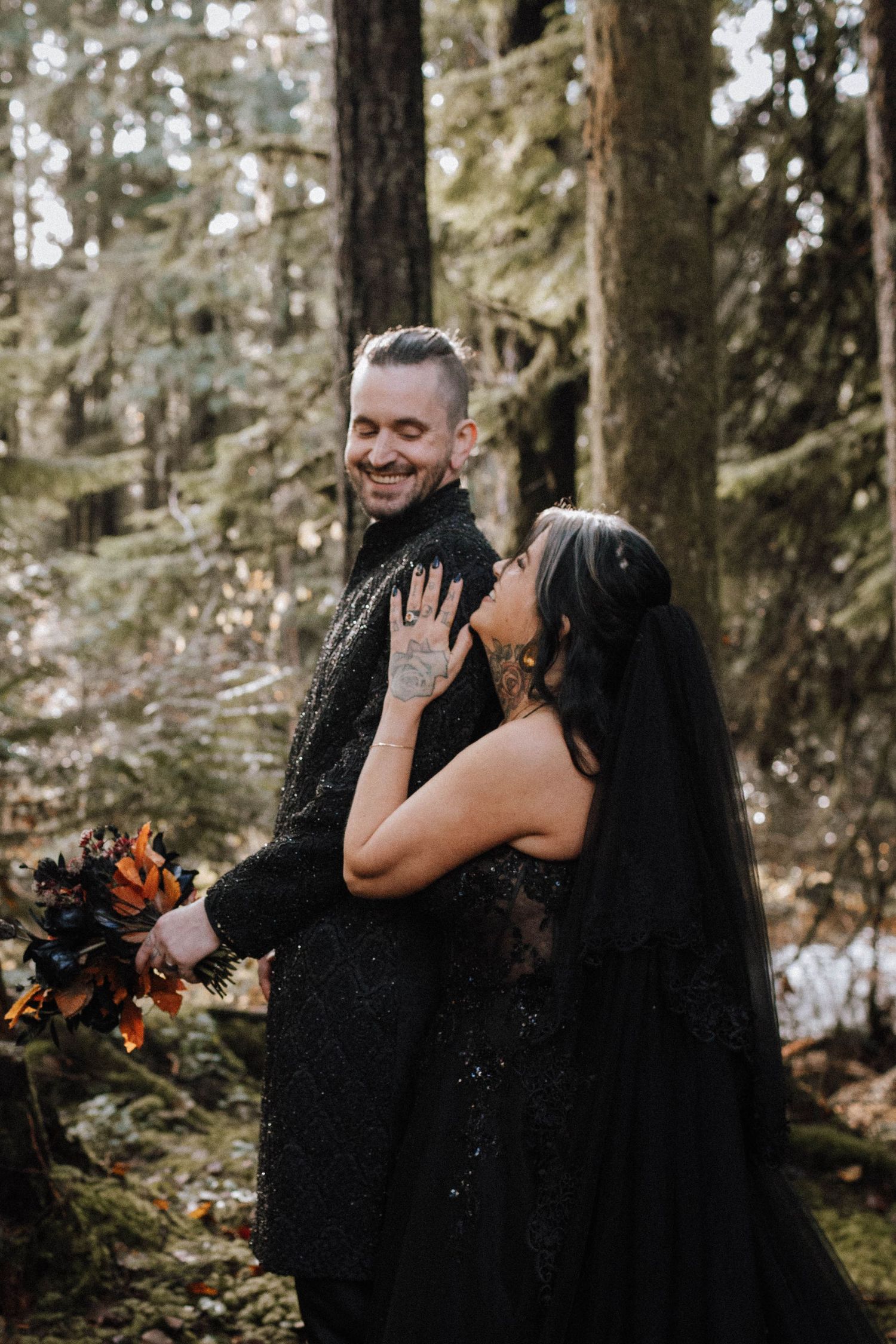 A happy couple in black formal wear shares a tender moment in a lush forest setting.