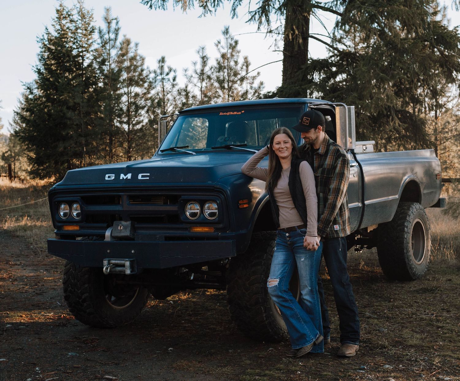 A couple stands beside a lifted classic GMC pickup truck surrounded by pine trees.