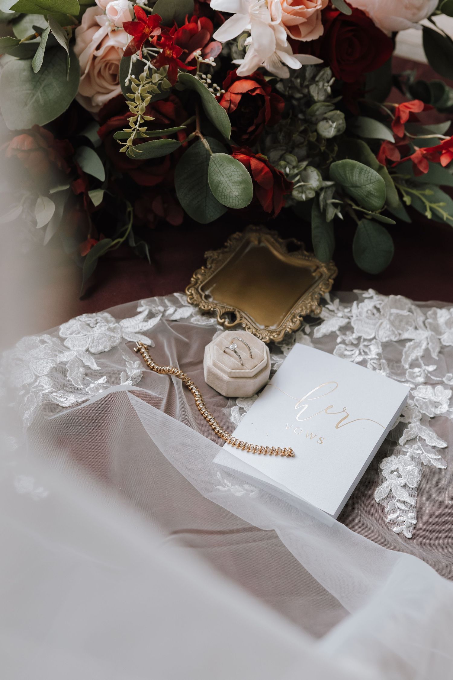 Wedding flat lay with lace veil, gold mirror, ring box, bracelet, and red and pink floral bouquet.