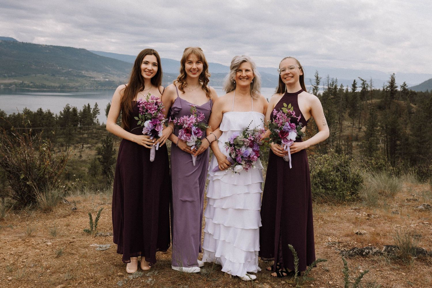 A bridal party wearing purple and lavender dresses poses together on a scenic cliff overlooking a lake and mountains.