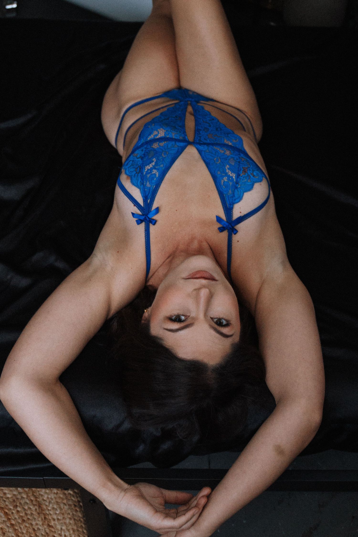 Person wearing blue lace lingerie poses on dark fabric while lying down.