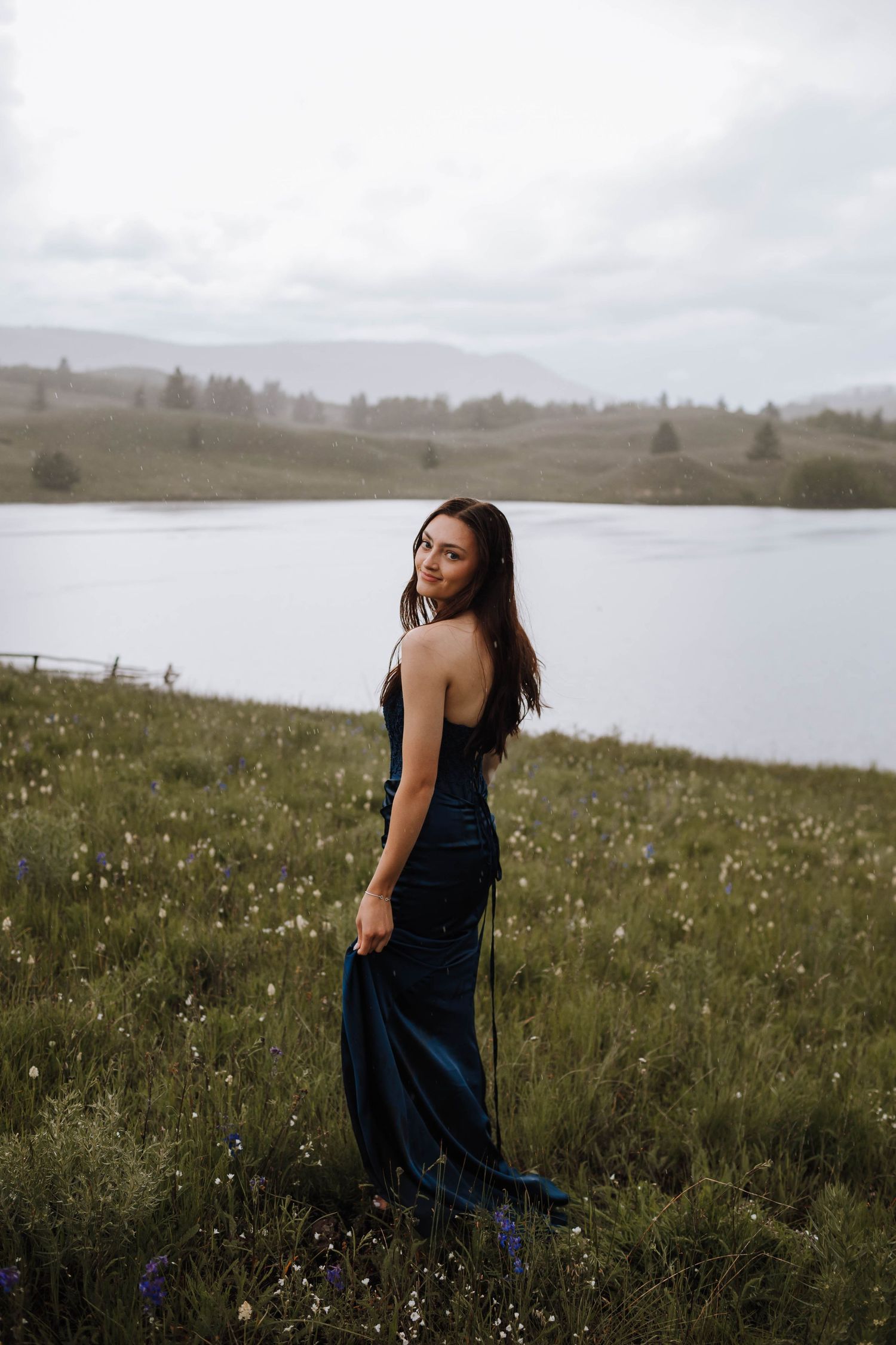 A navy blue dress contrasts against wildflowers and a serene mountain lake backdrop.