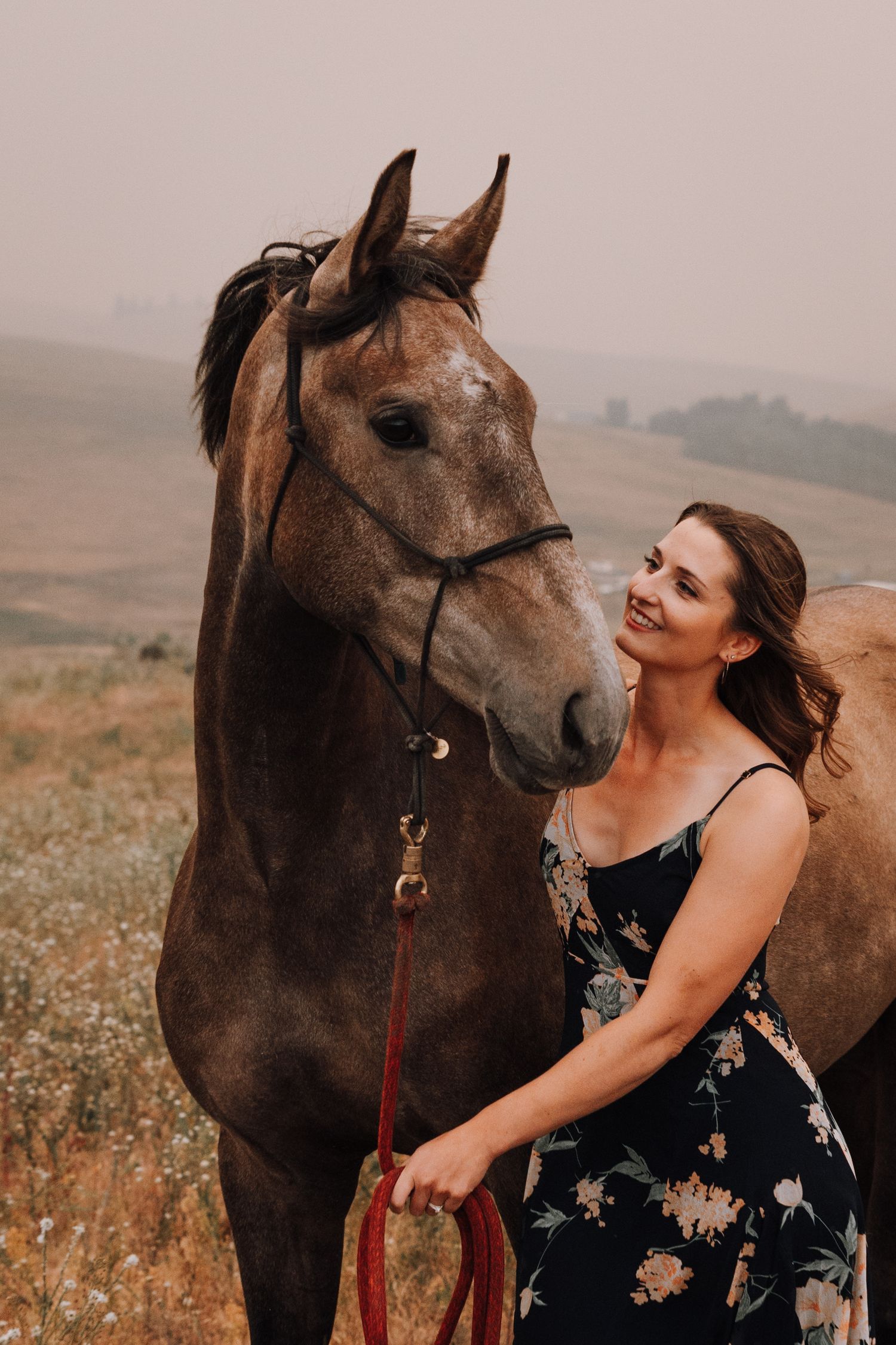 A smiling woman in a floral dress holds the reins of a brown horse in a misty field.