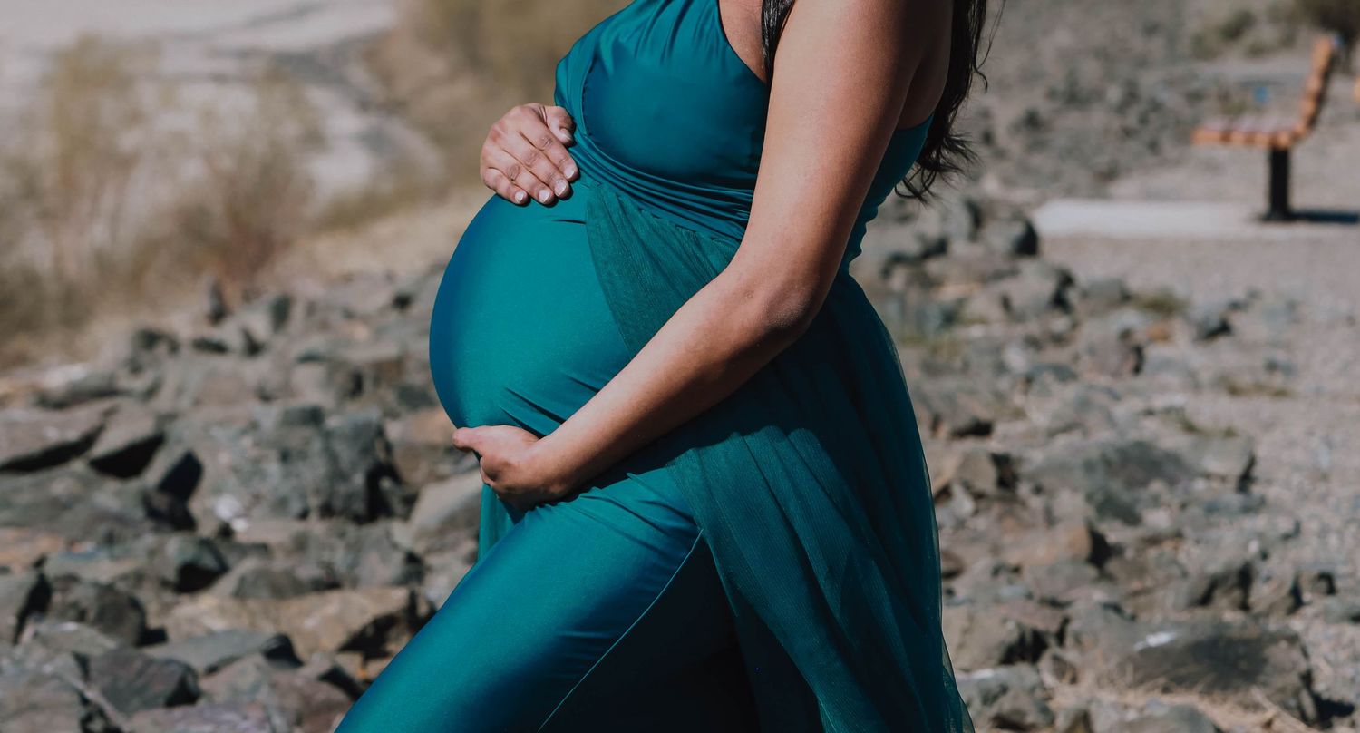 An elegant teal maternity dress is showcased against a rocky outdoor backdrop.