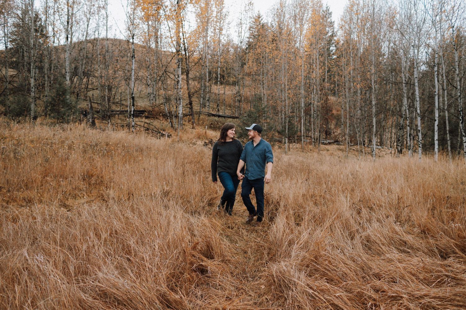 Couple walking through tall autumn grass with bare trees in background.