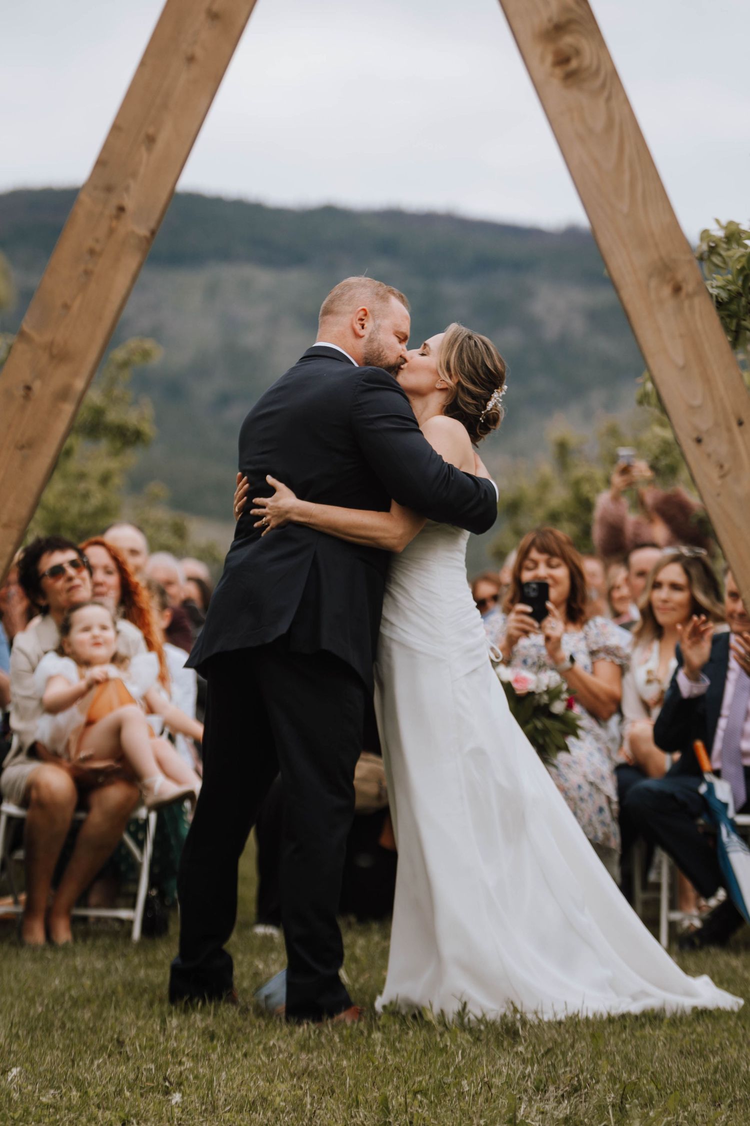 A couple shares their first kiss as newlyweds under a wooden arch while wedding guests look on from their seats.
