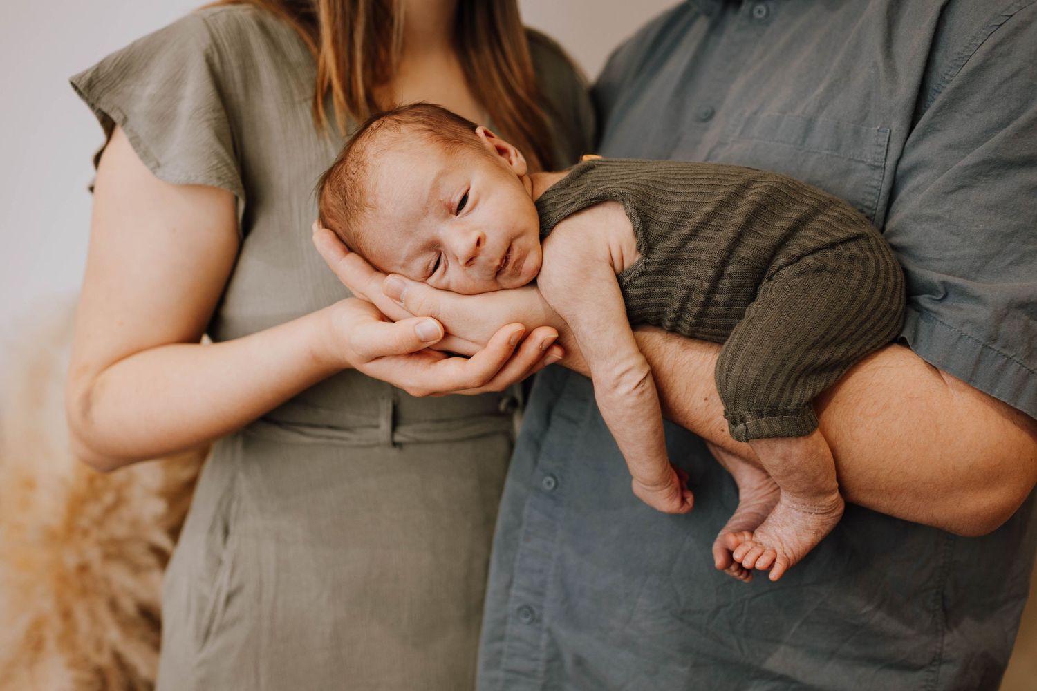 A sleeping baby in dark gray overalls rests peacefully between two adults in neutral colored clothing.