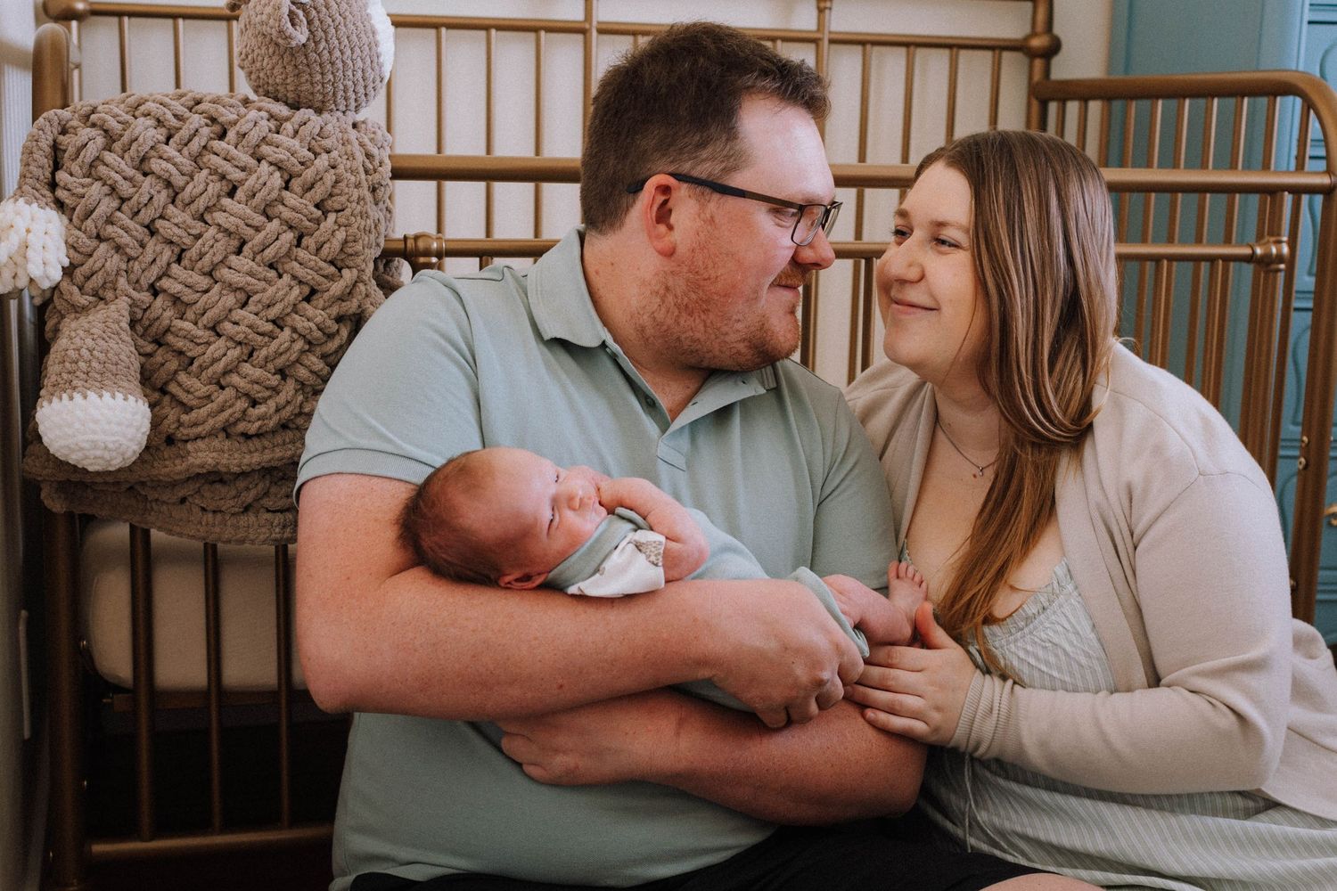 A loving couple sits together in a nursery holding their newborn baby near a wooden crib and macrame wall decor.