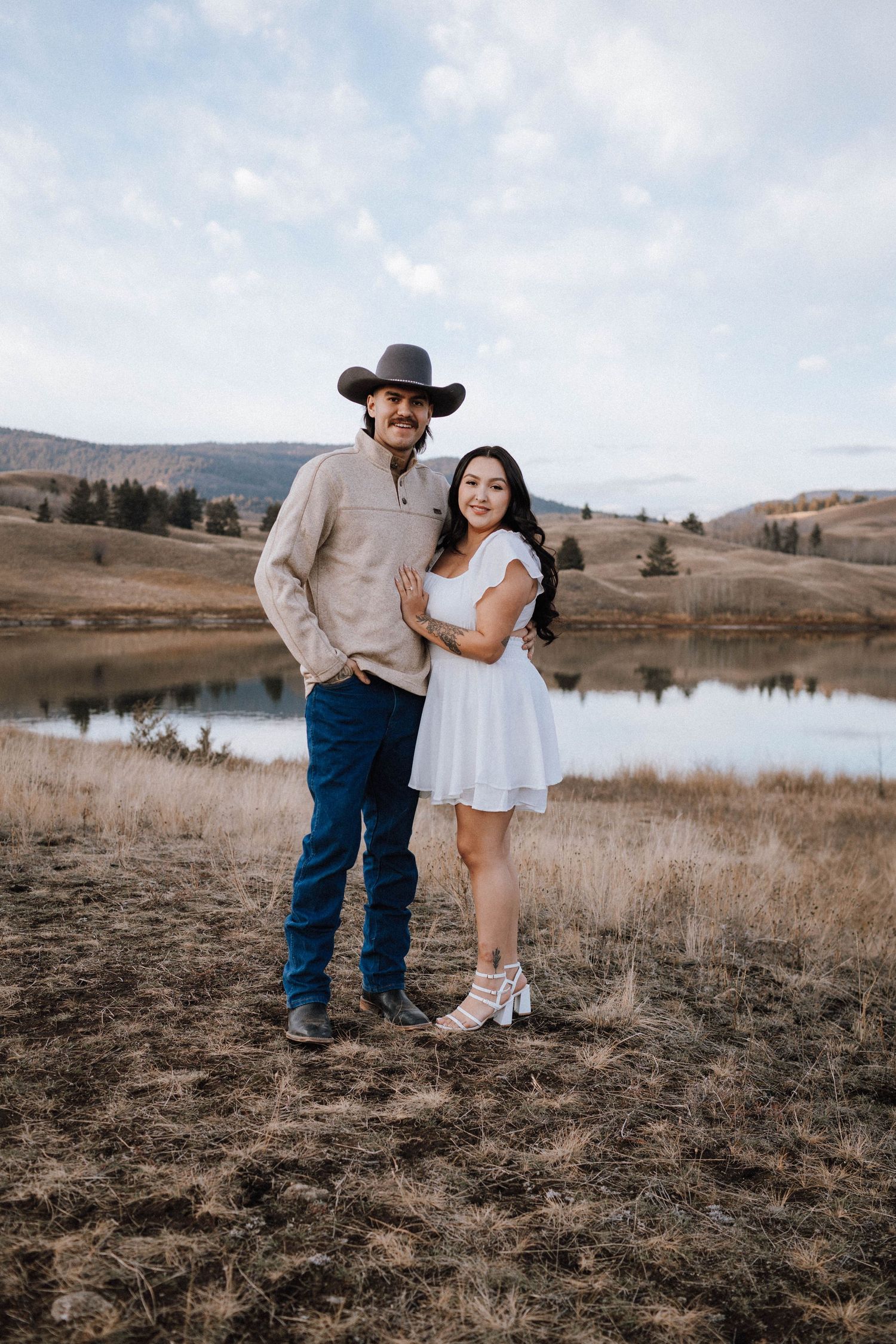 A couple in western attire poses together on a hillside overlooking a serene lake during a photo session.
