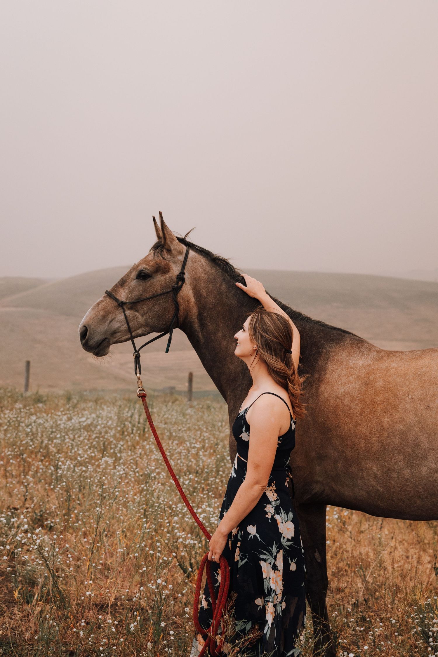 Woman in floral dress tenderly stroking a brown horse in a misty field.