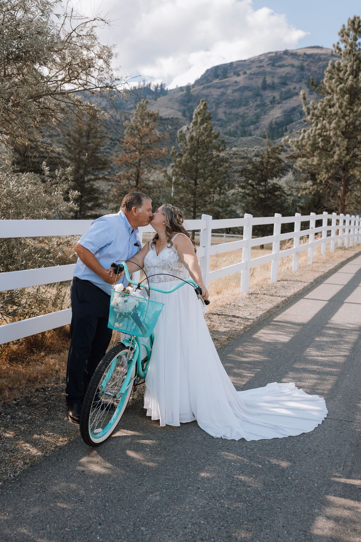 Bride in white gown kisses groom by teal bicycle on scenic mountain path with white fence.
