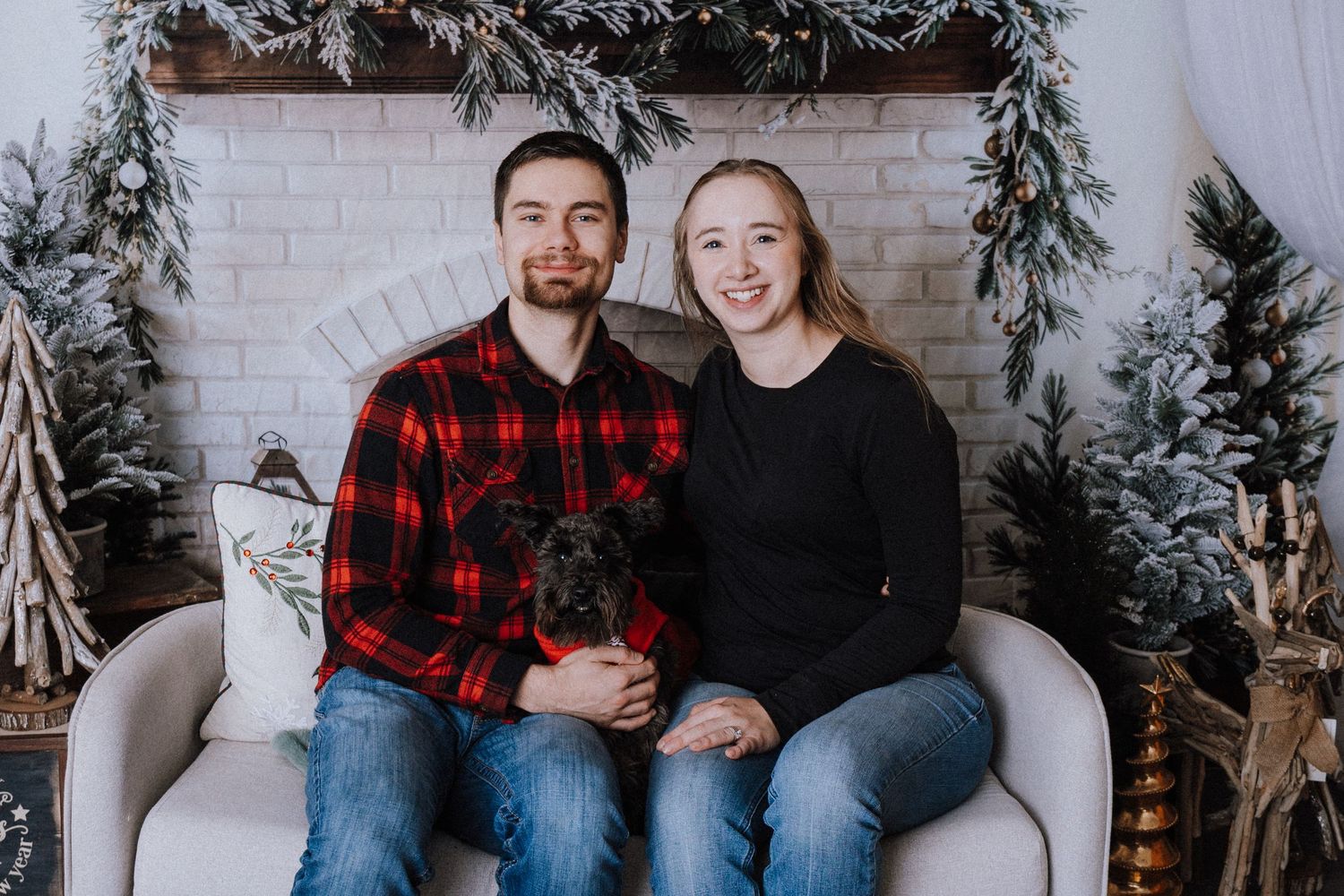Couple sitting on white chair with small black dog in front of Christmas fireplace decor with snowy trees.