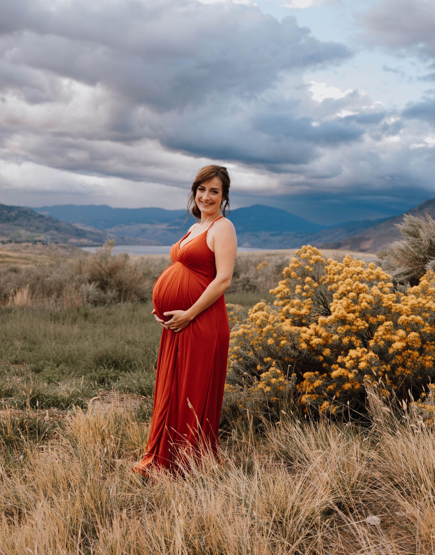Expectant mother in flowing red dress poses in mountain landscape with stormy sky and yellow flowers.