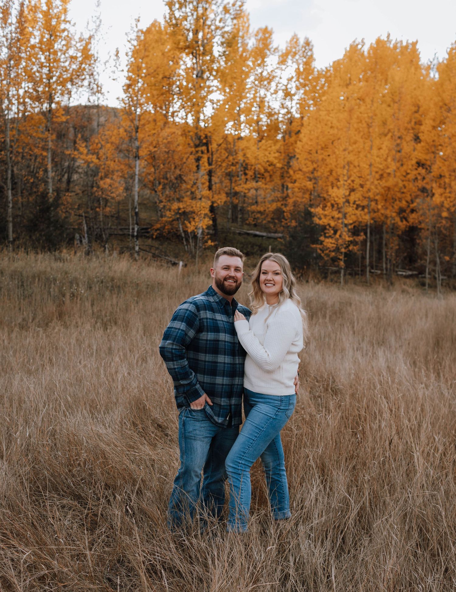 Couple stands together in tall grass field with golden autumn aspens in background.