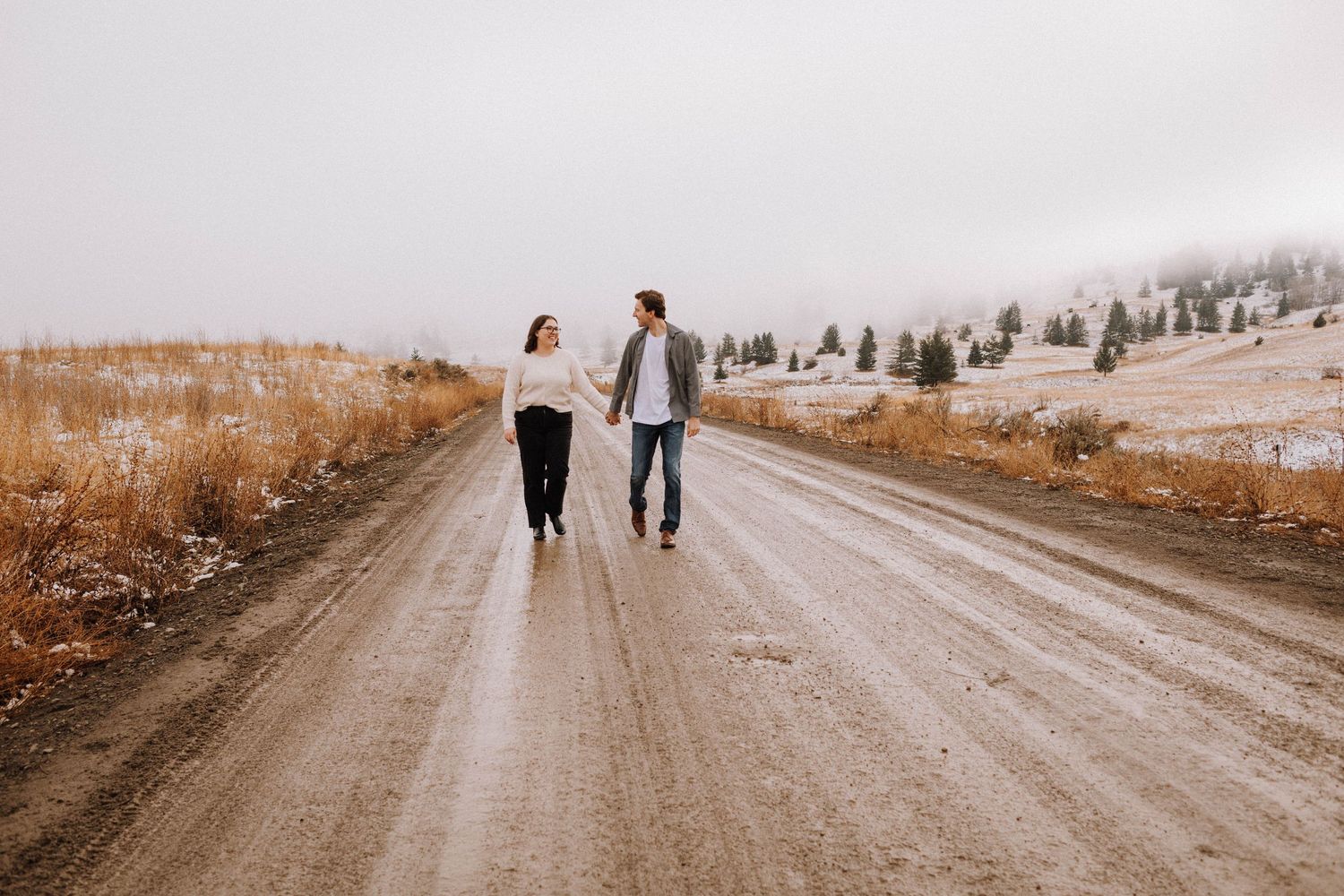 A couple walks hand in hand down a dirt path surrounded by autumn colored grass and trees in the distance.