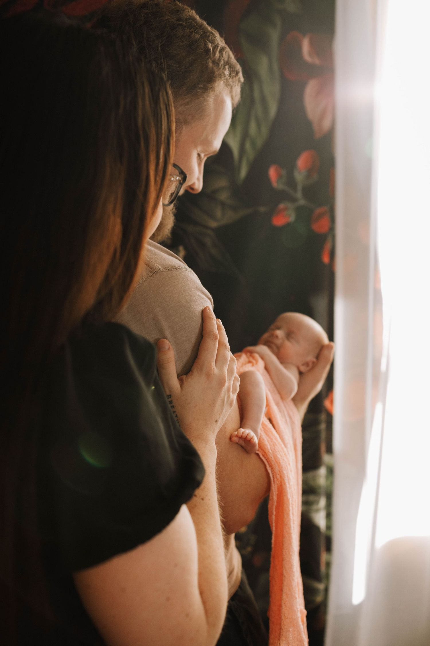 Tender moment between parent and infant in soft window light with floral backdrop.