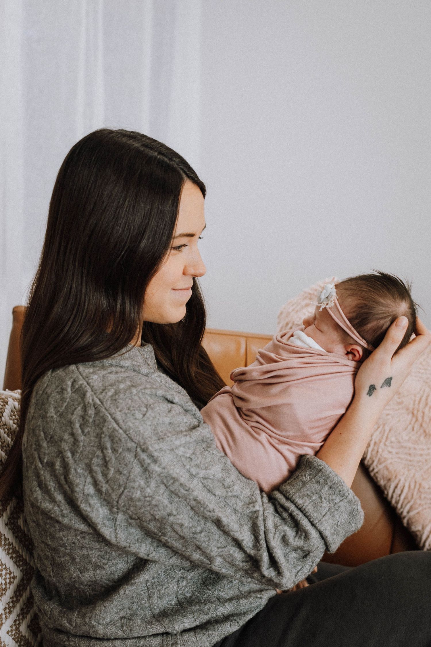 A tender moment between parent and newborn baby wrapped in pink, both wearing cozy sweaters on a leather couch.