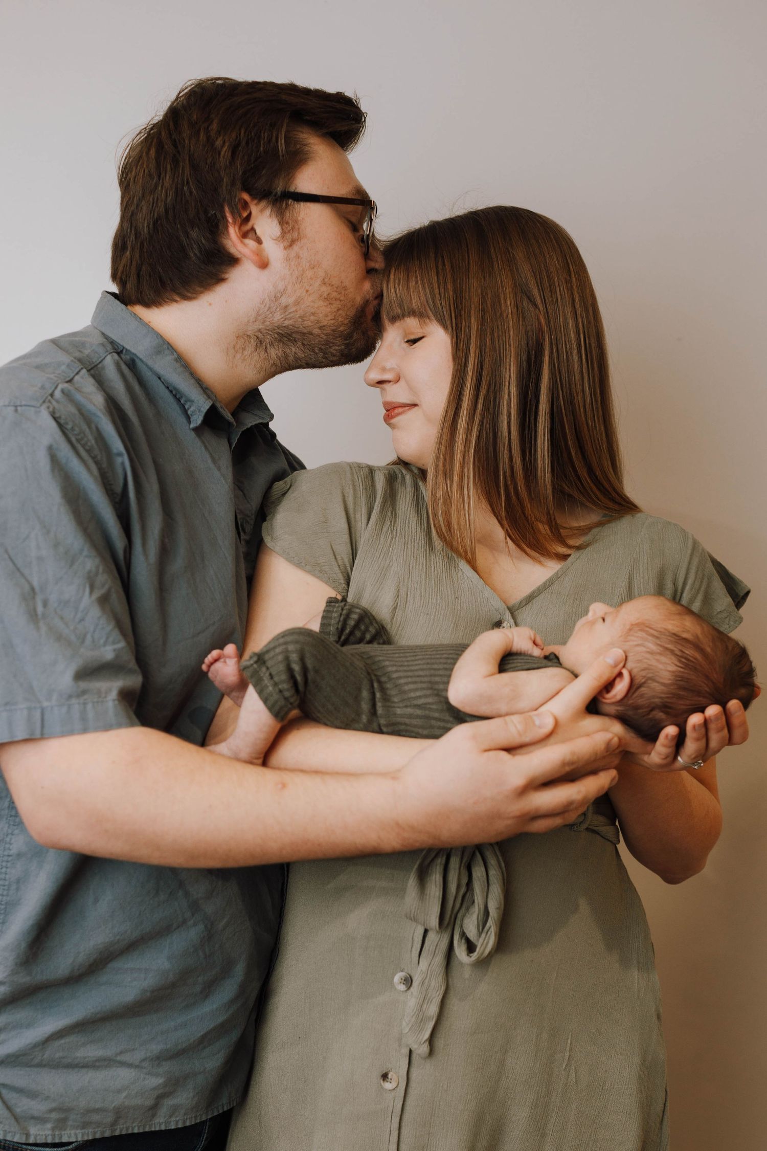 A tender family moment as parents embrace while holding their newborn baby against a neutral background.