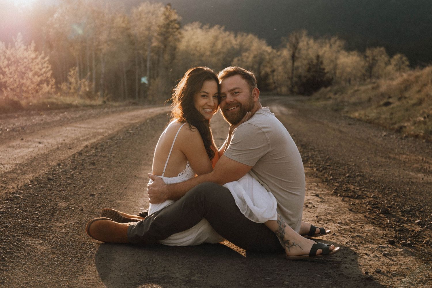 A couple sits intimately together on a dirt road at sunset with warm golden light illuminating the scene.