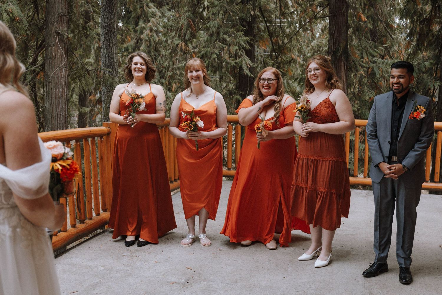 A wedding party in rust-colored dresses and dark suits poses outdoors among tall trees in a forest setting.