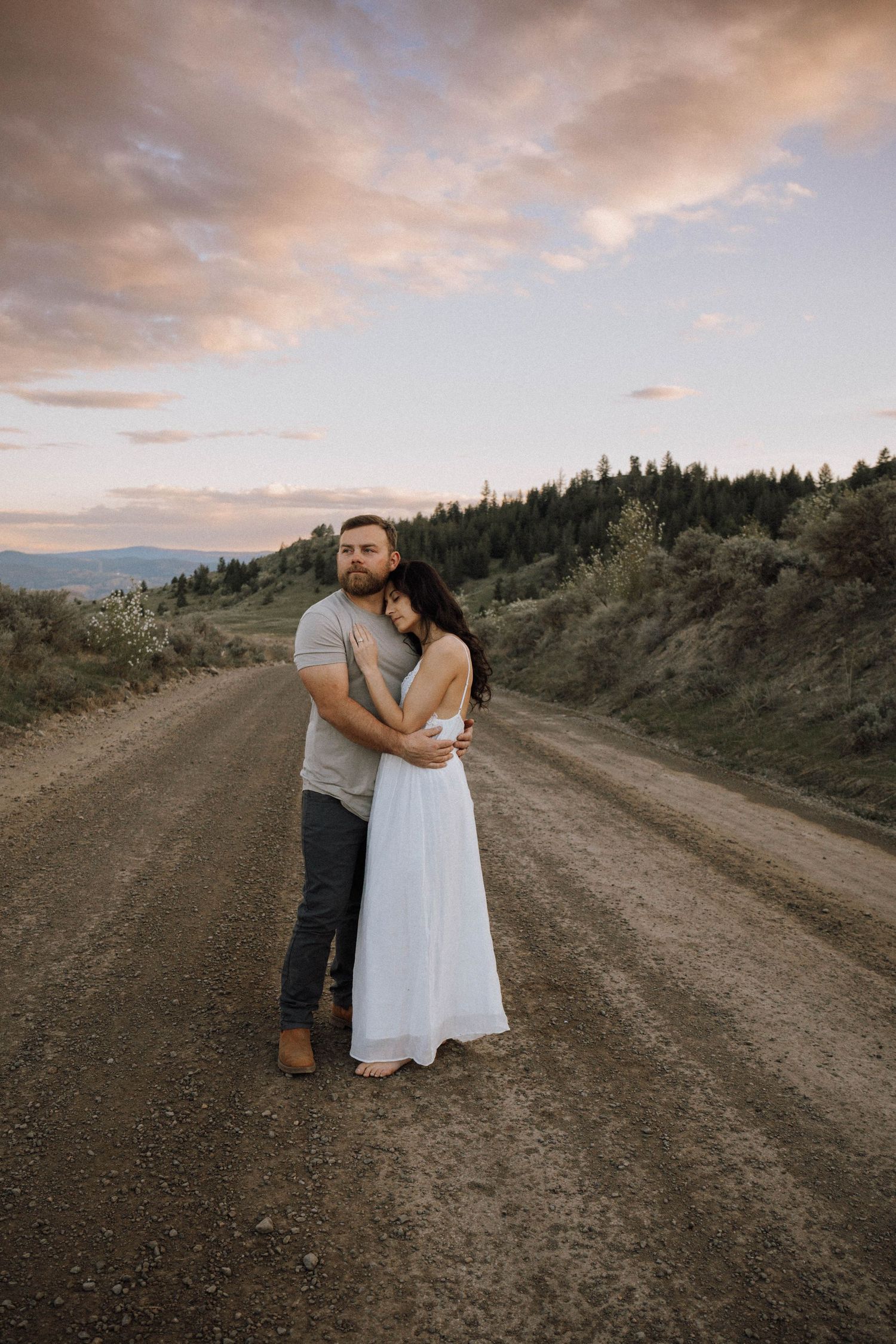 A couple embraces on a dirt mountain road at sunset with evergreen forest and pink clouds in the background.