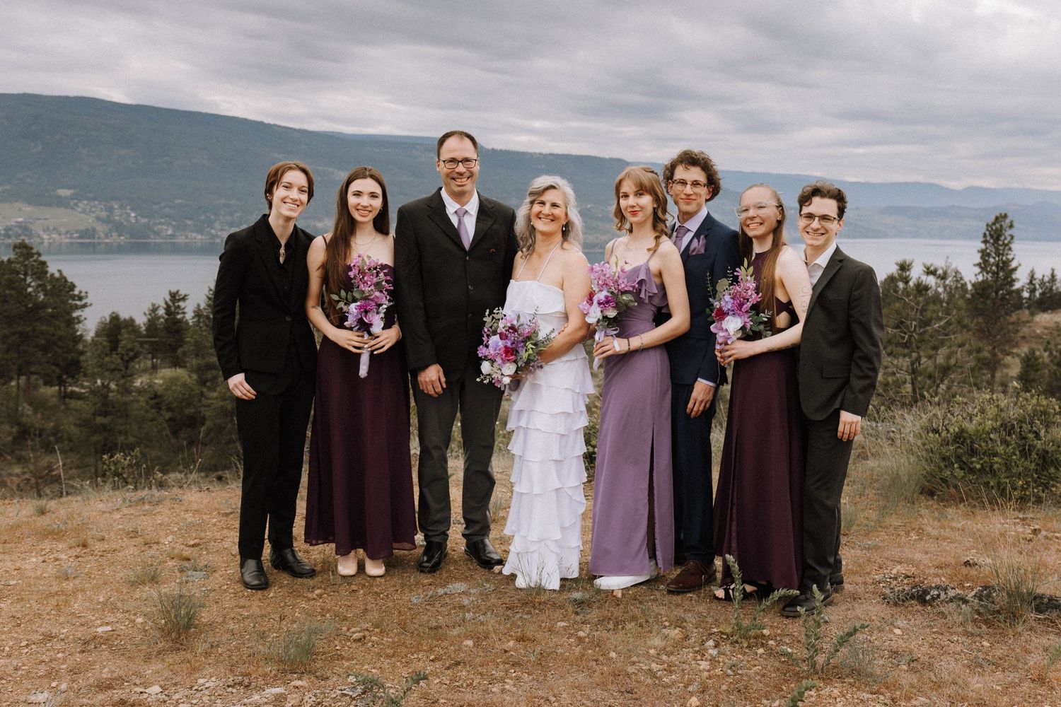 Wedding group photo featuring bridal party in burgundy and purple dresses against scenic mountain landscape.