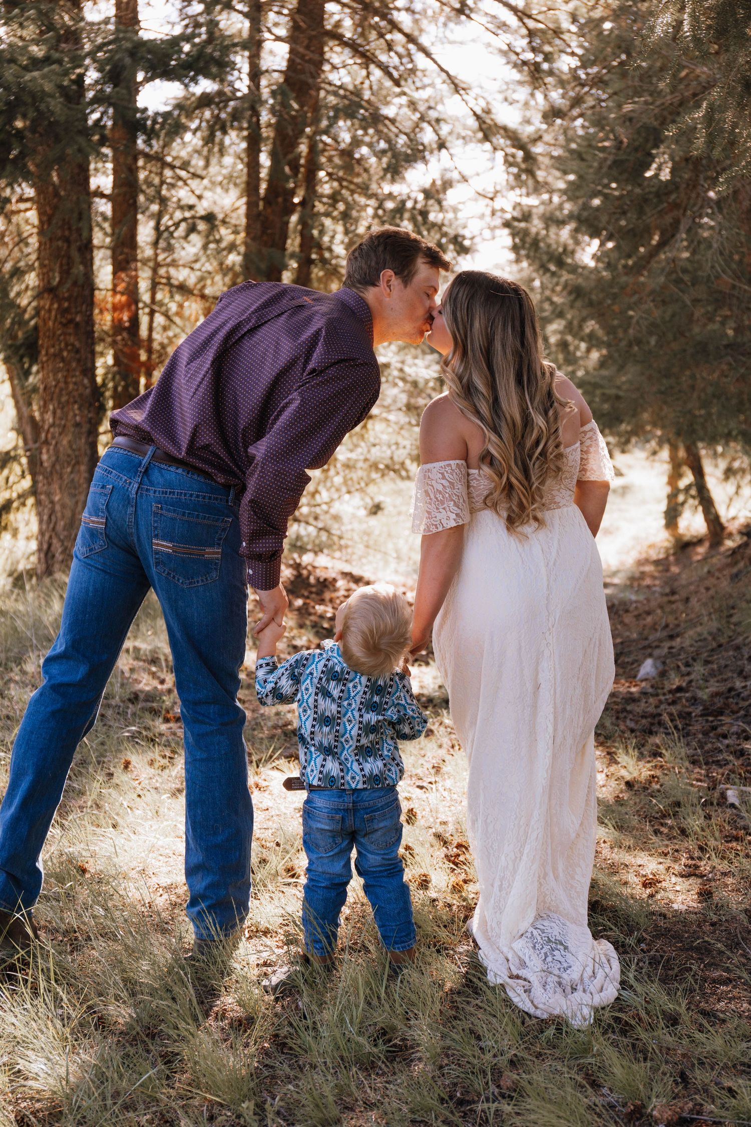 A couple and toddler share a tender moment outdoors among pine trees at sunset during a family photo session.