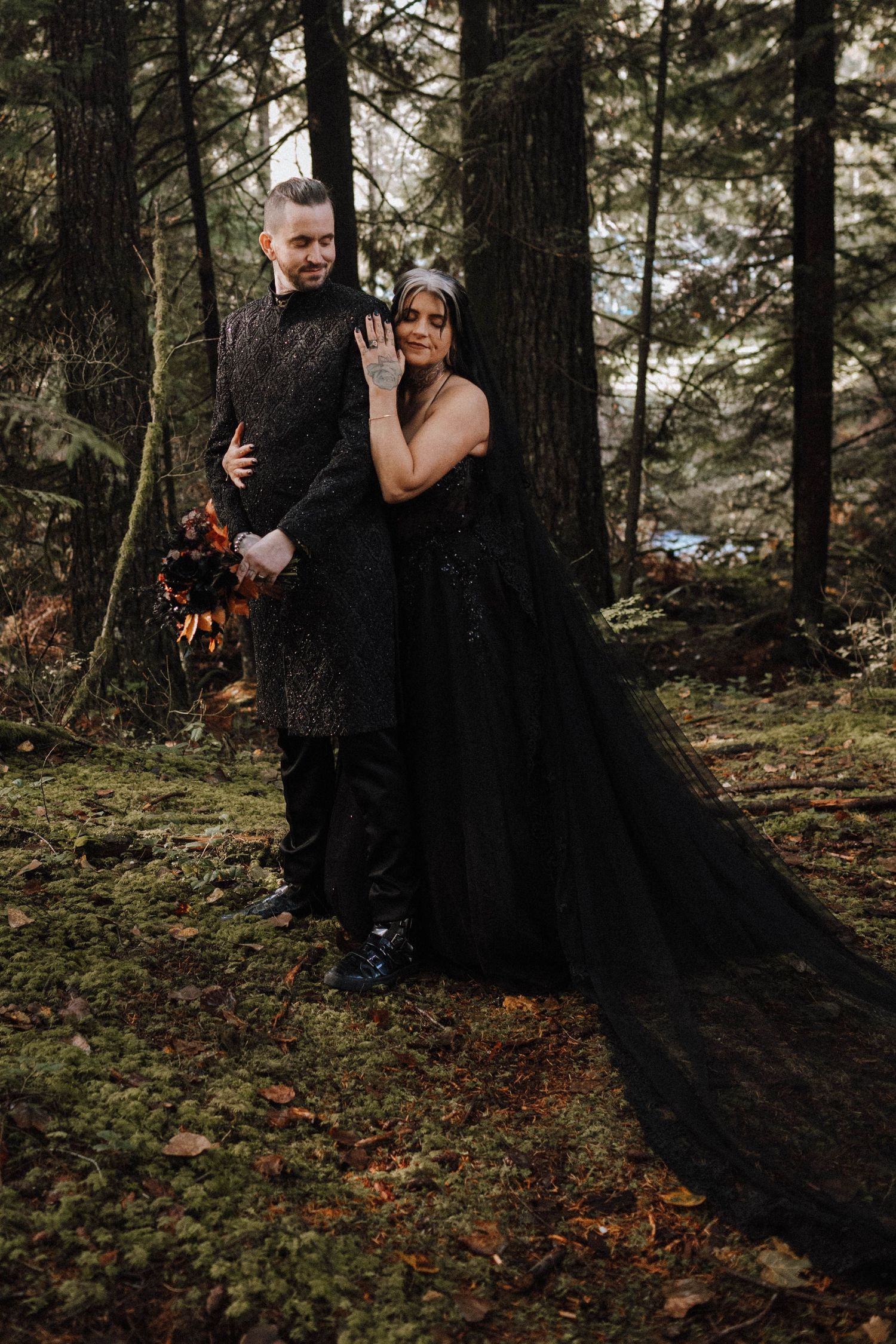 A couple poses in a moody forest setting wearing black formal attire with a dramatic flowing black dress.