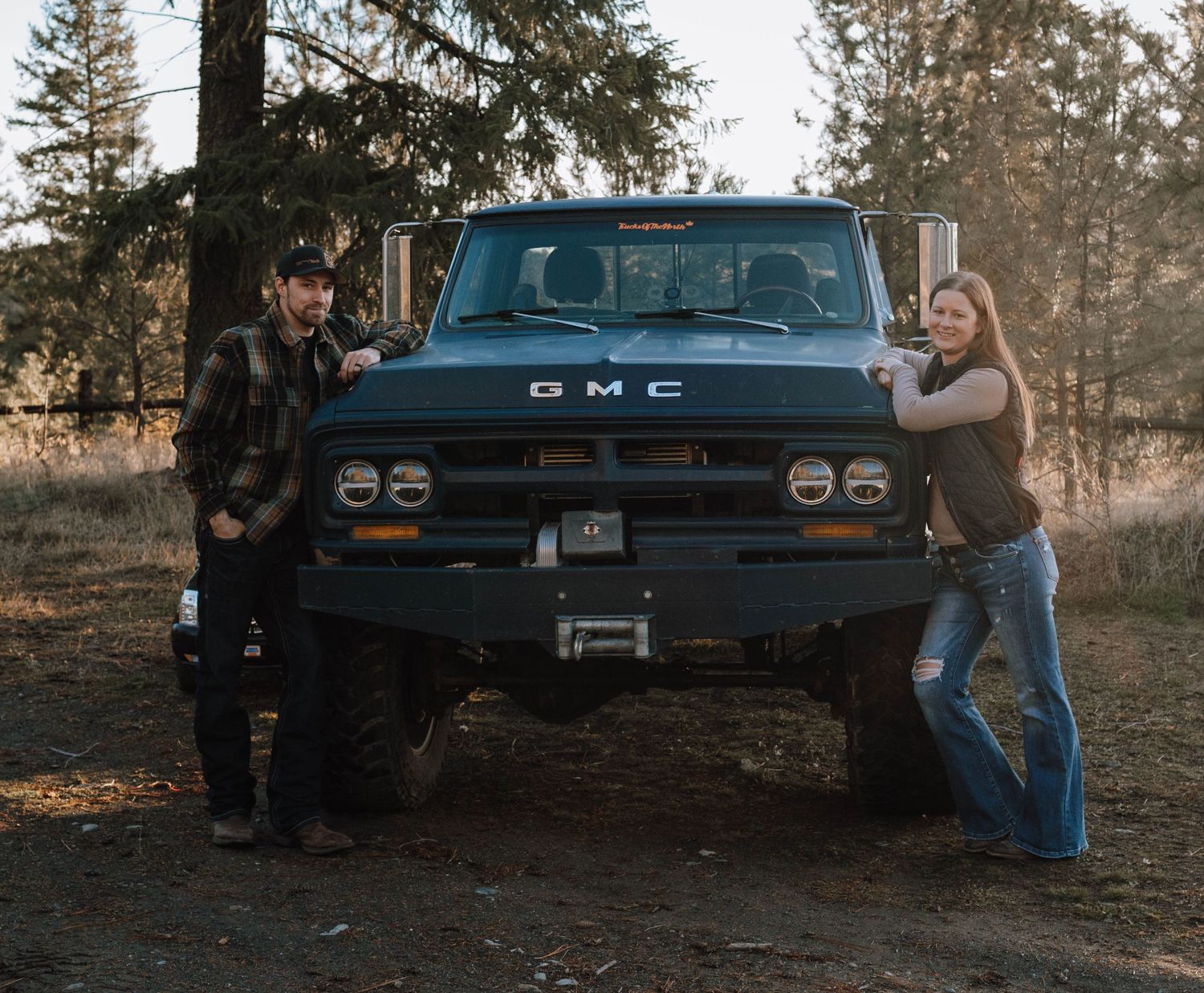 A couple poses with their modified vintage GMC truck in a forest setting.