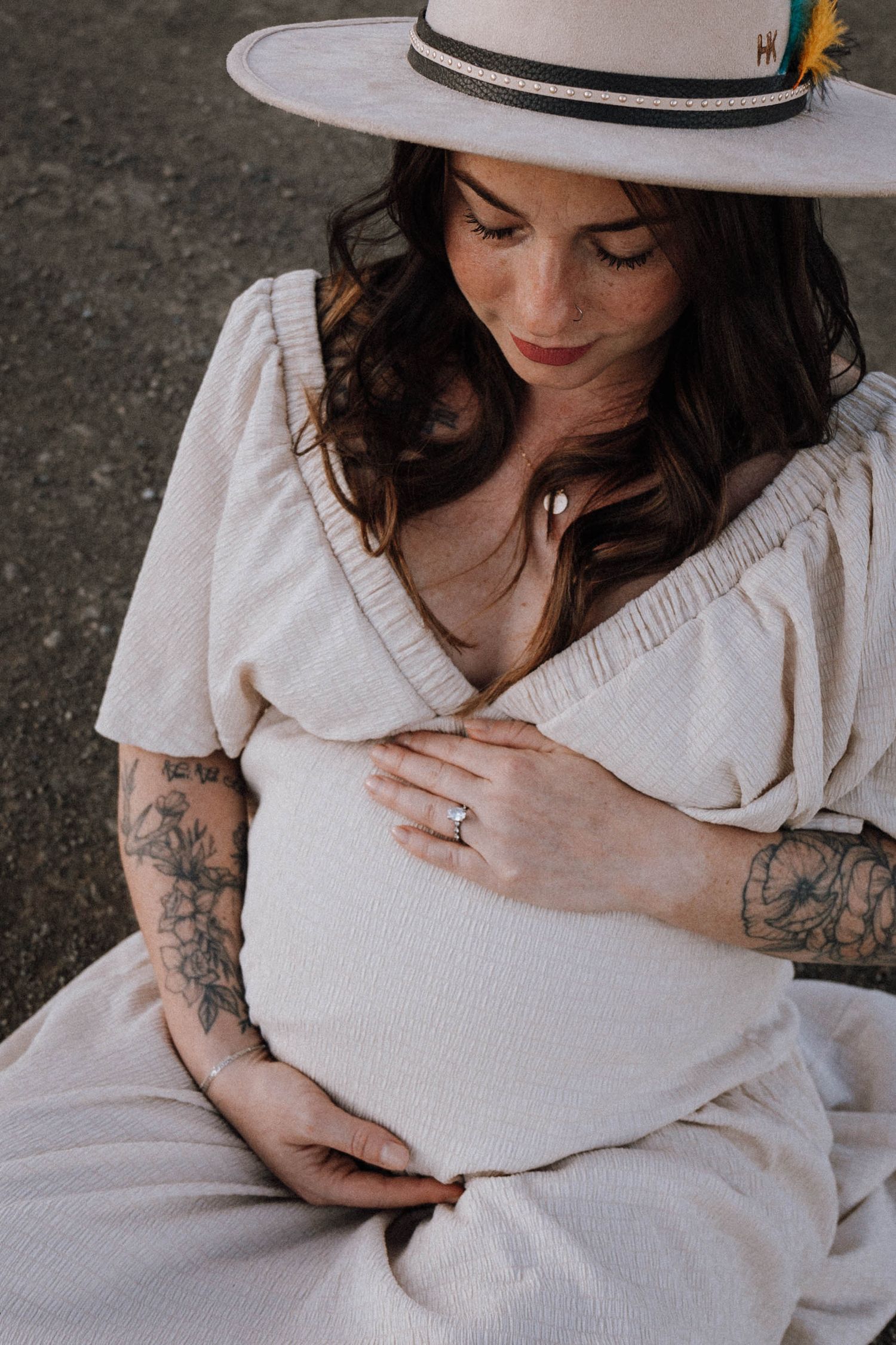 Artistic maternity photo series showing woman in white dress and hat touching her pregnant belly against grey backdrop.