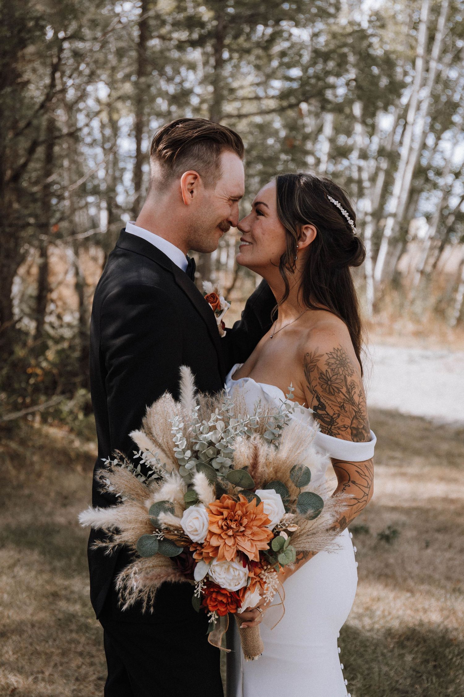 Romantic wedding couple embraces in a birch forest with autumn-colored bouquet and off-shoulder white dress.