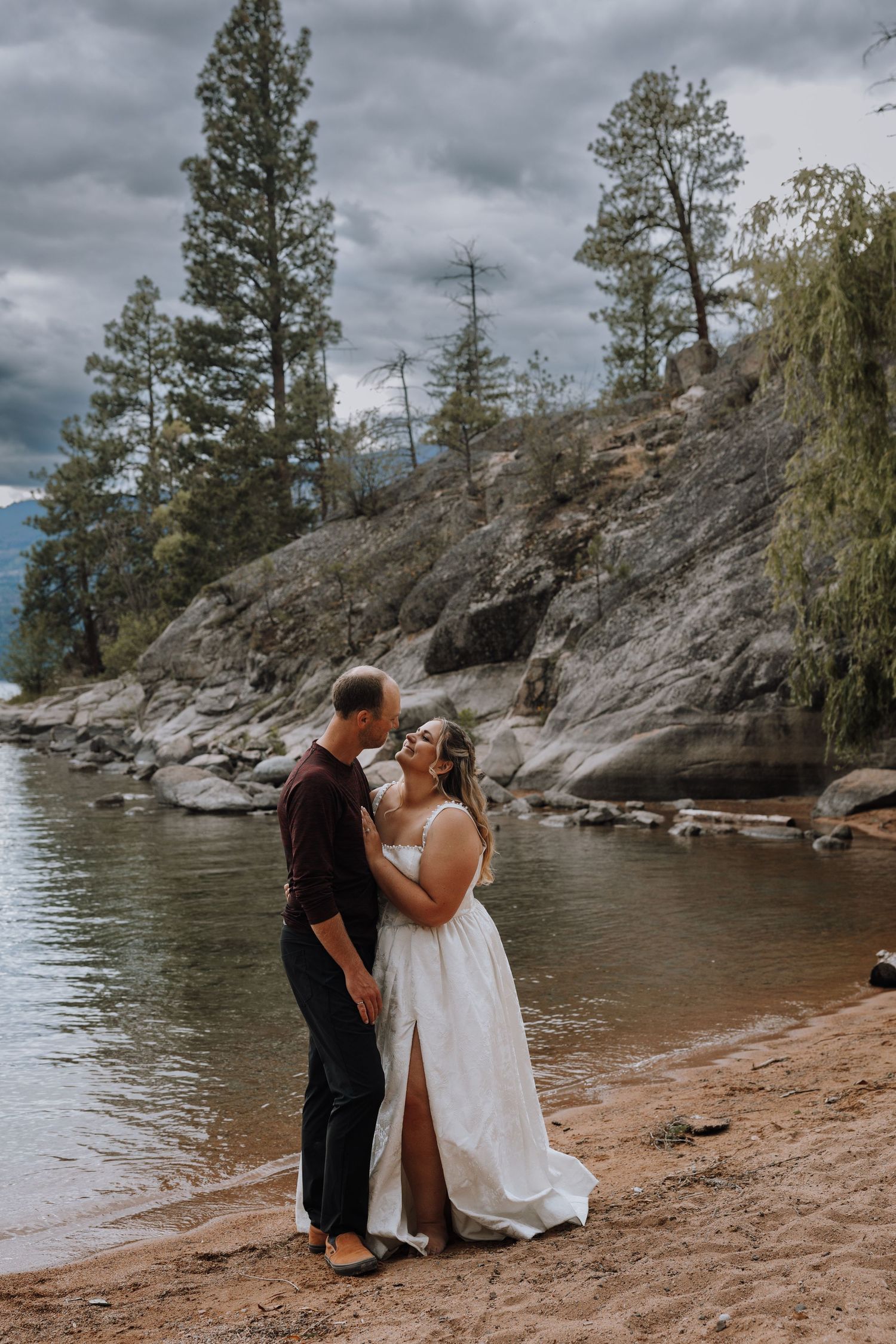 Couple shares romantic moment on sandy lakeside beach, bride in white gown, groom in dark attire, pine trees backdrop.