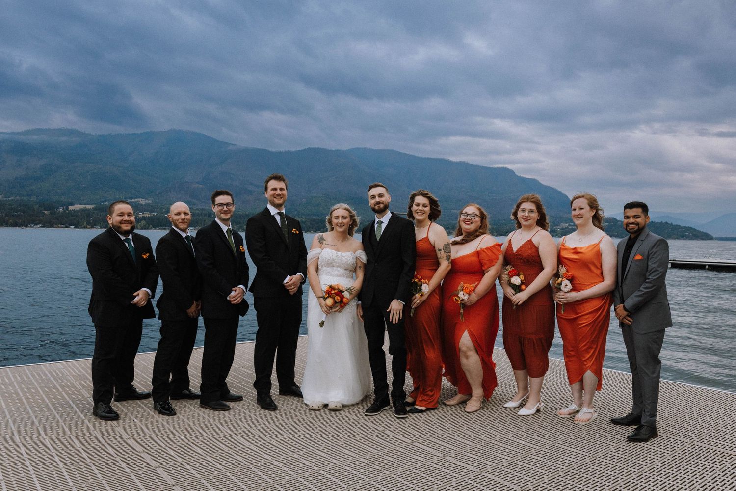 Wedding party poses for panoramic photos against dramatic mountain and lake backdrop at sunset.
