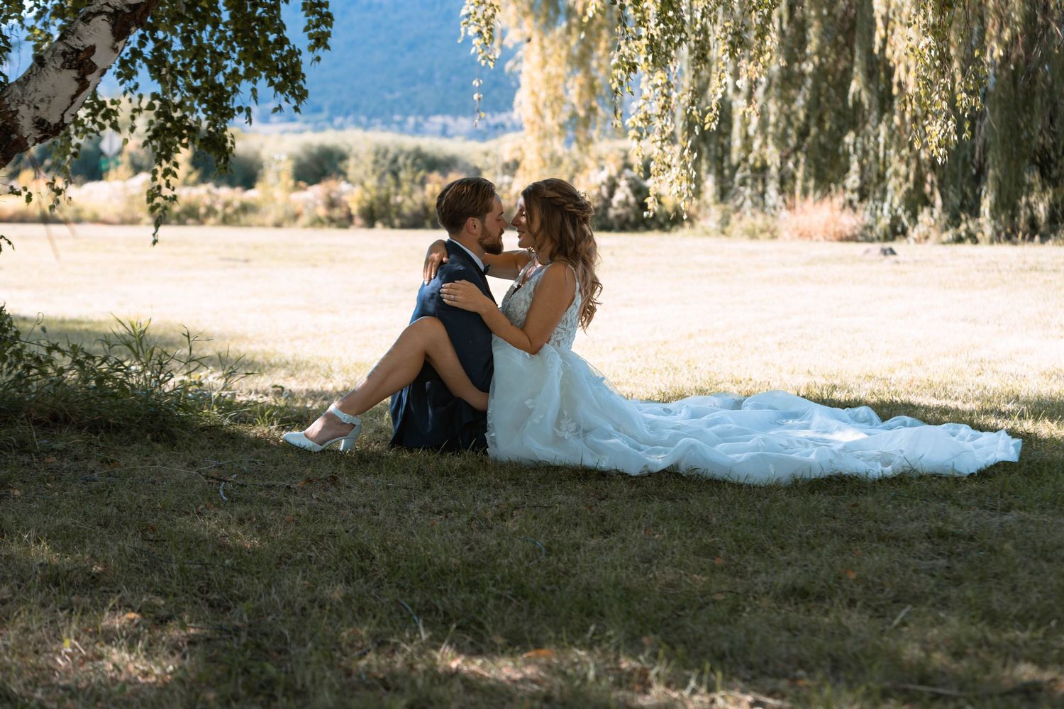 Couple relaxes under a willow tree on their wedding day, with the bride wearing a flowing white dress.