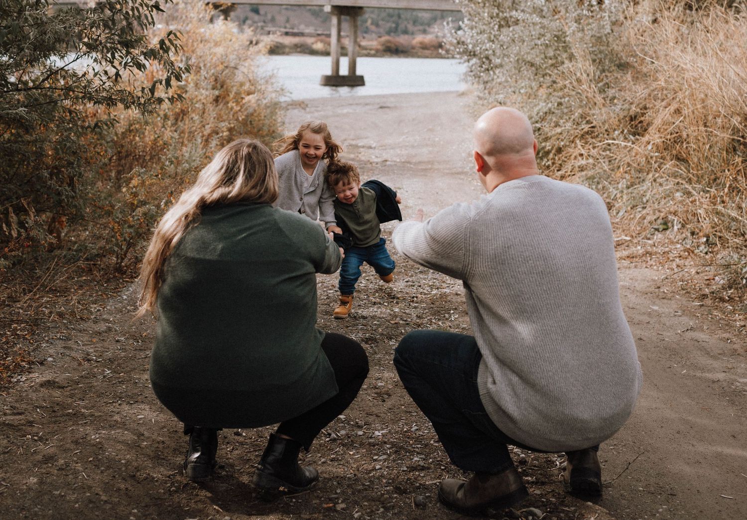 Two adults crouching and playing with a small child on a dirt path surrounded by autumn foliage.