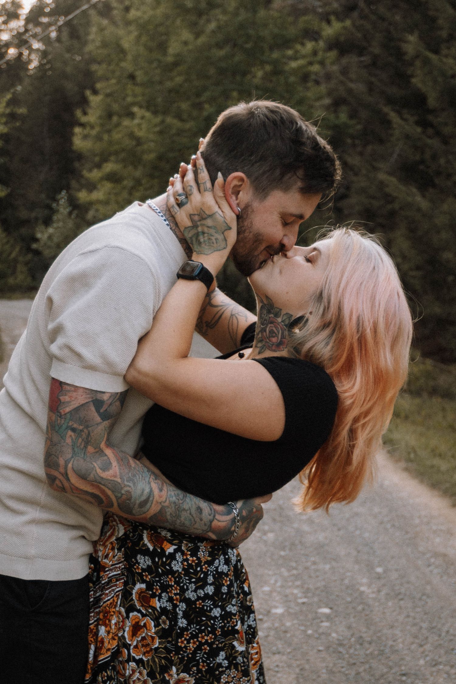 A romantic couple embraces on a forest path, with one person wearing a floral skirt and the other showing colorful arm tattoos.