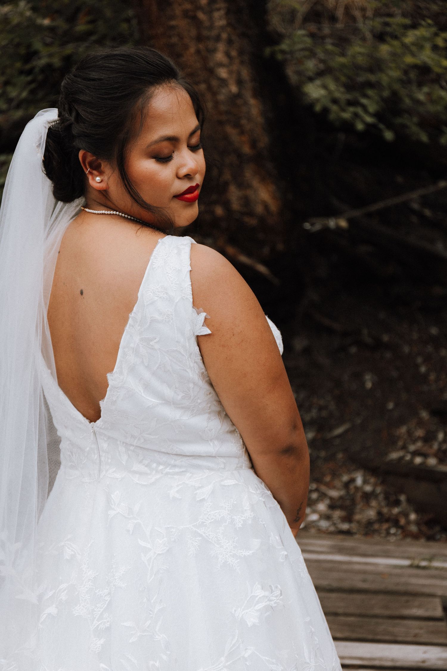 A bride in a white lace wedding dress poses in an outdoor setting with a flowing veil trailing behind her.