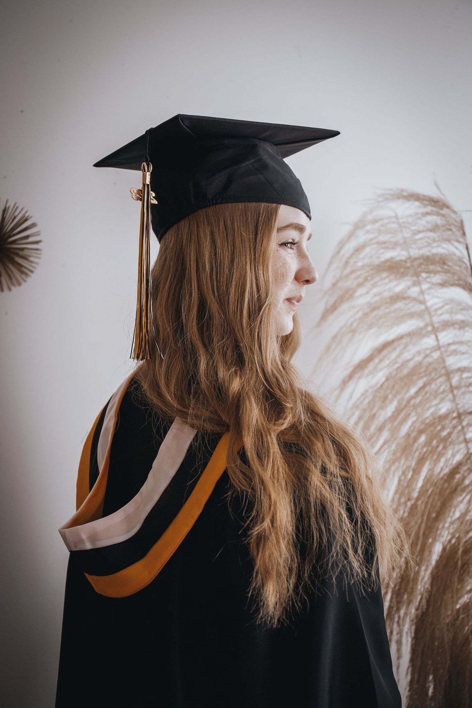 Graduate in cap and gown poses against white wall with pampas grass decoration.