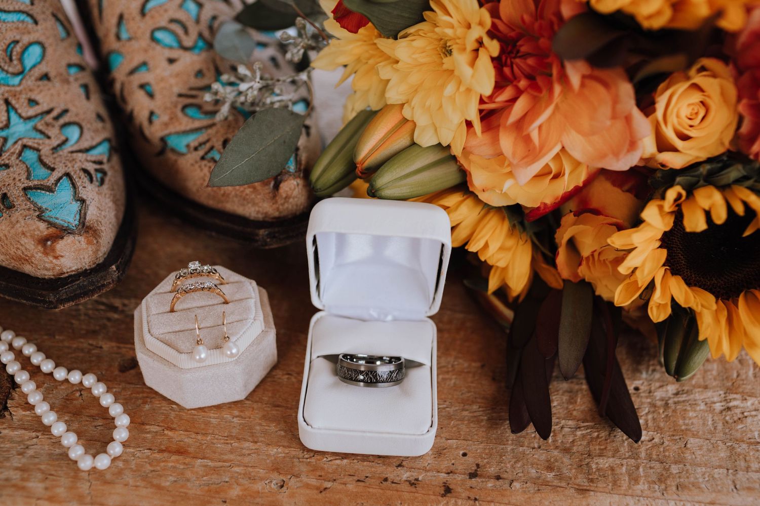 Wedding ring in white box with orange sunflowers and cowboy boots on wooden surface.