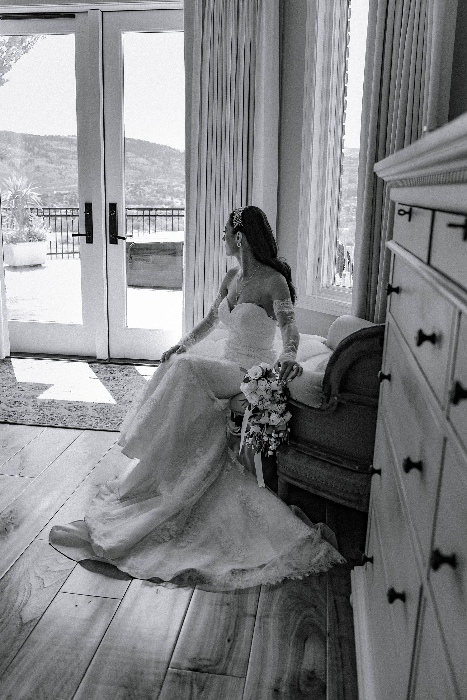Black and white photo of a bride in an elegant wedding gown sitting by a window with mountain views and natural light.