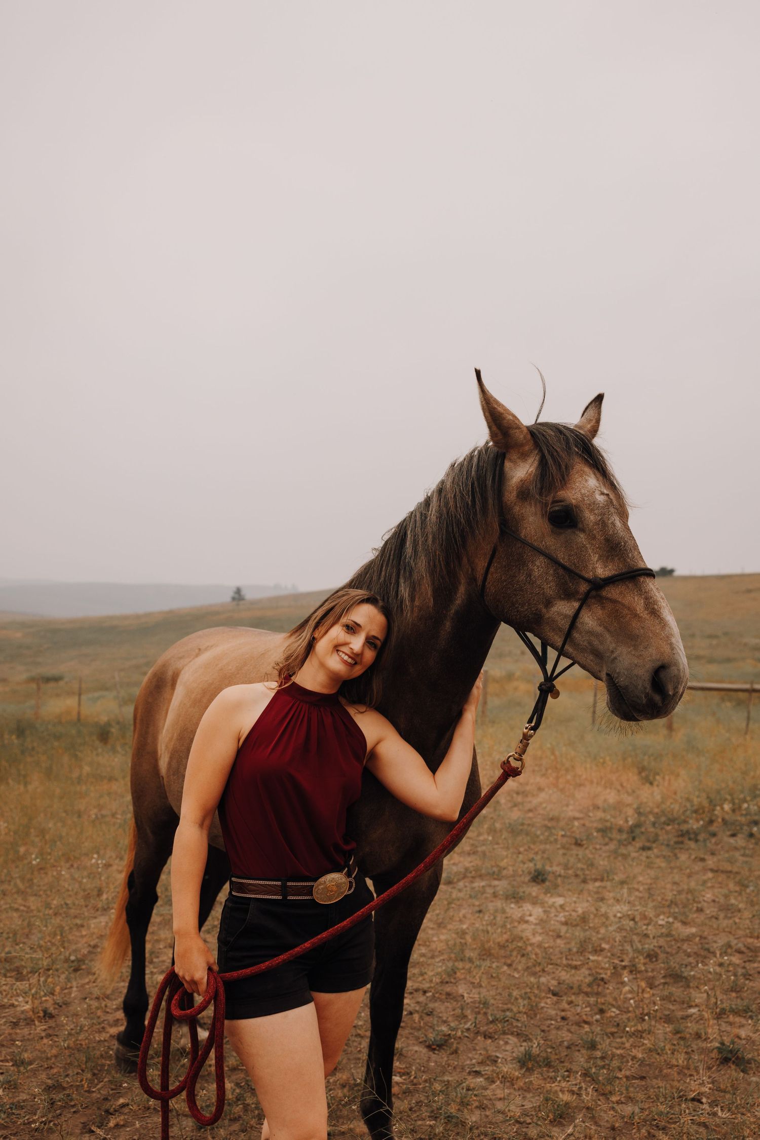 Smiling woman in burgundy top and black shorts stands beside a brown horse on a grassy rural landscape.