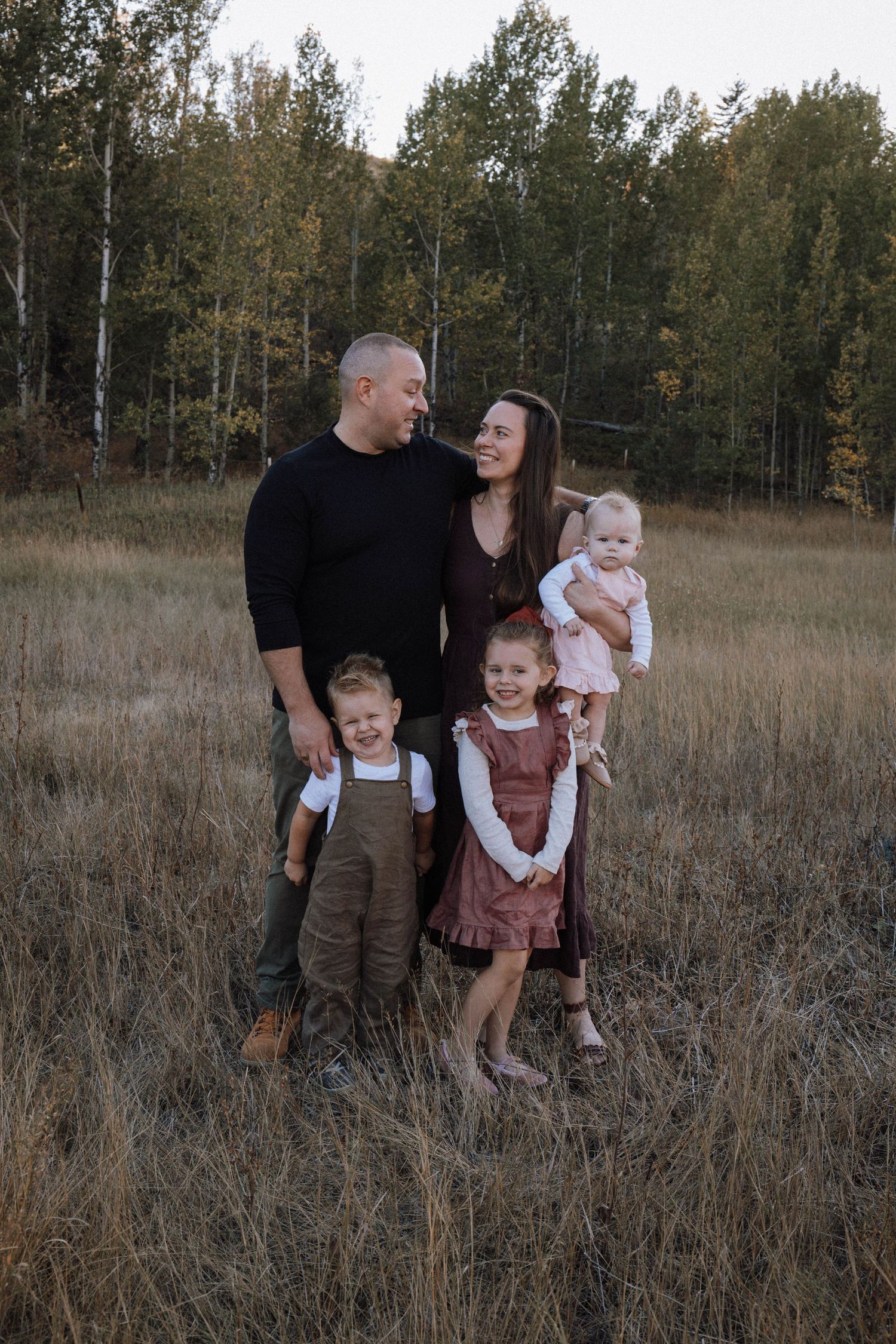Family stands together in tall autumn grass with trees in background during golden hour outdoor photo session.