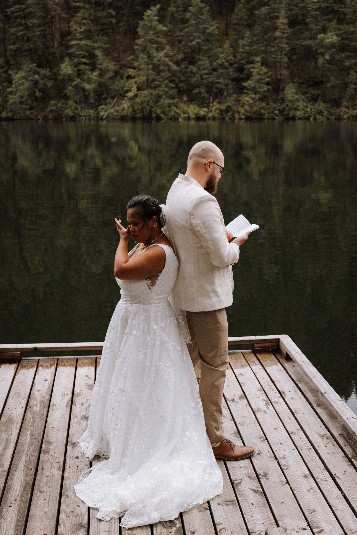 A couple in wedding attire shares a playful moment on a wooden dock by a serene lake surrounded by evergreen trees.