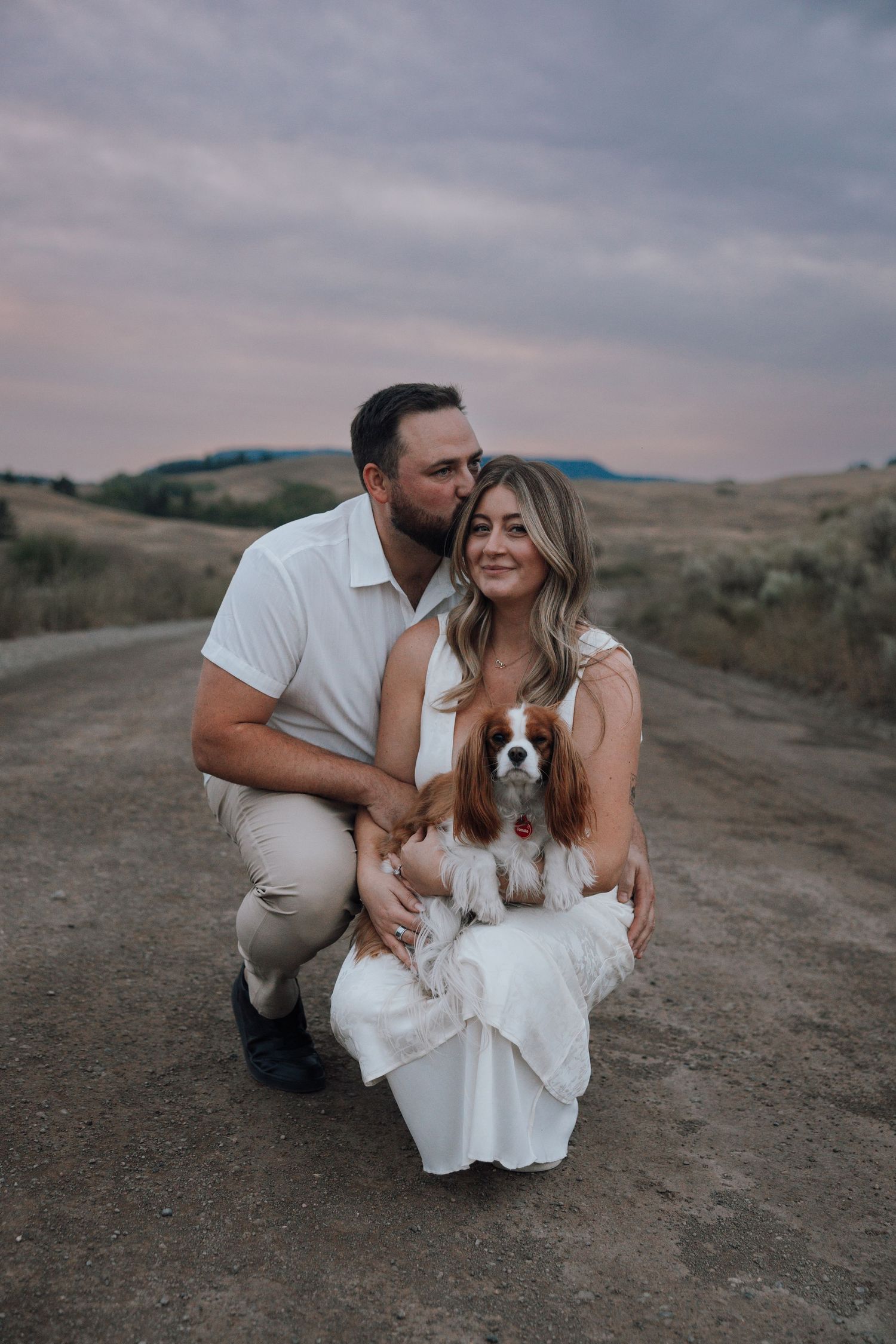Couple crouching on a dirt road at dusk, man kissing woman's cheek while holding a Cavalier King Charles Spaniel.