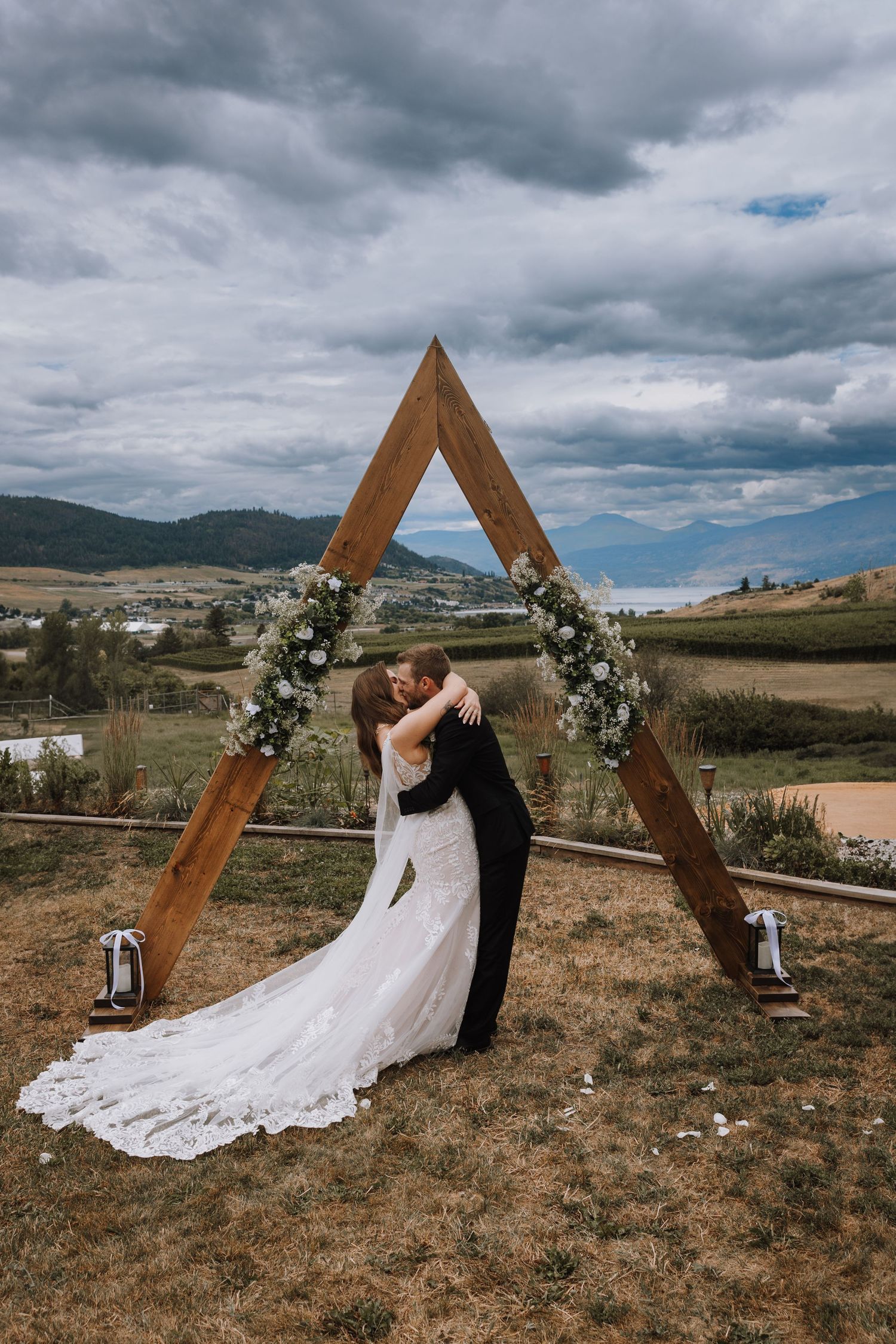 Bride and groom share first kiss under a triangular wooden arch adorned with white flowers, set against a scenic mountain backdrop.