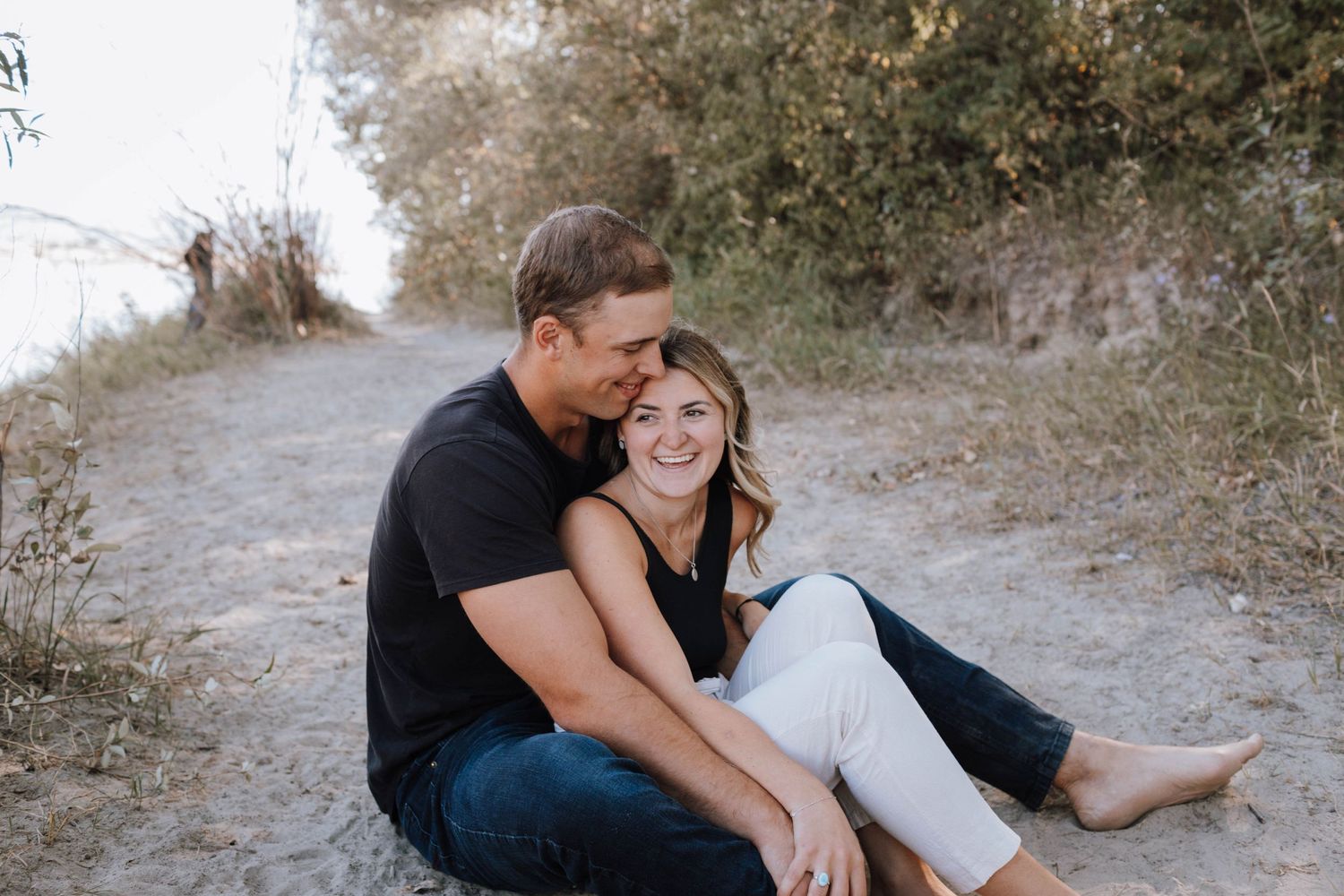 A couple shares an intimate moment while sitting on a dirt trail by the water.