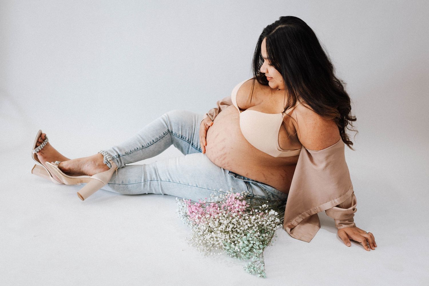 Artistic maternity photo with light pink lingerie, grey leg warmers, and delicate baby's breath flowers on white background.