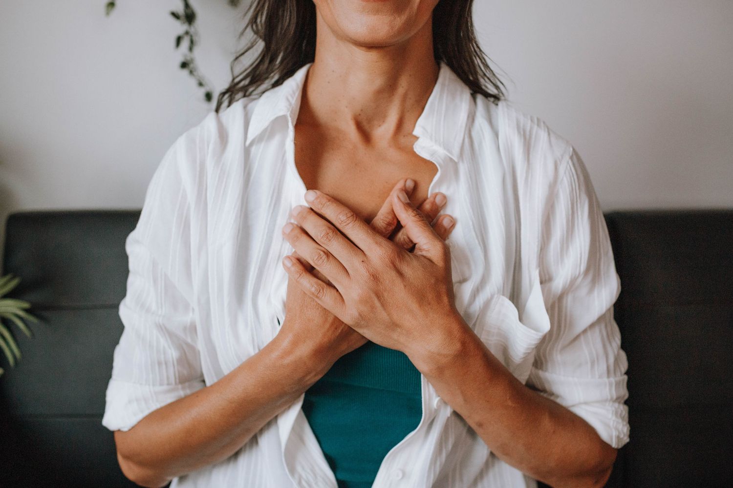 A minimalist indoor portrait showing hands clasped over heart against a dark sofa backdrop.