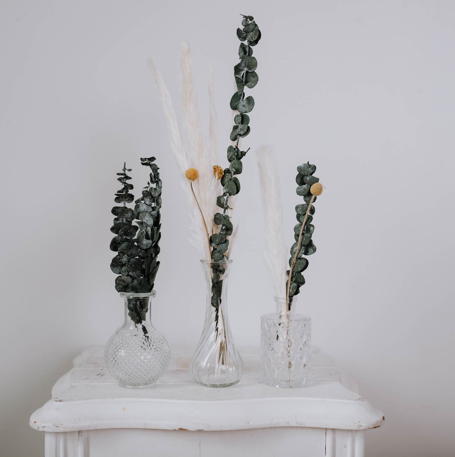 Three glass vases with eucalyptus stems and dried flowers arranged on a white table against a neutral wall.