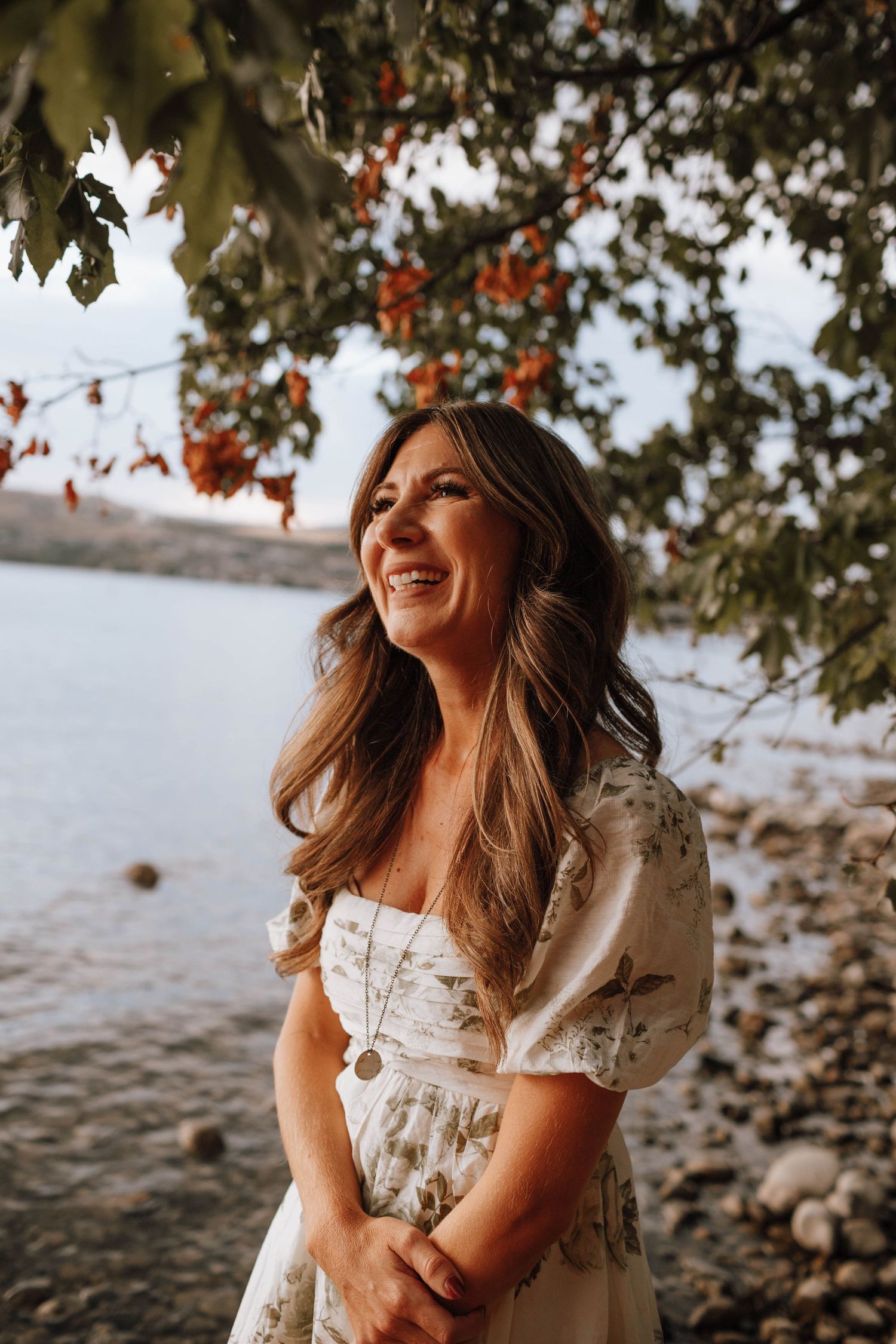 A flowing white dress catches the light near a lakeside with autumn trees in the background.